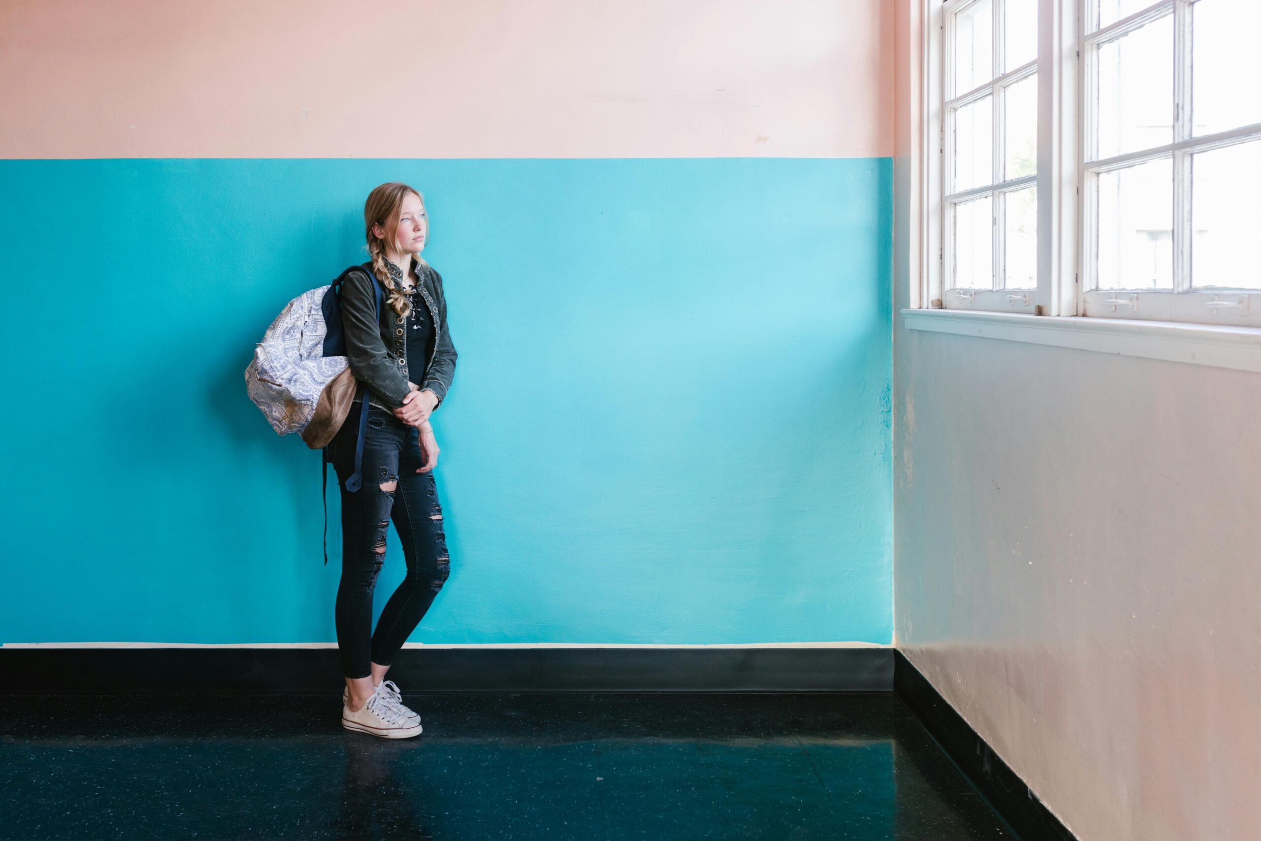 Teen student in casual attire with backpack looking out a window at school.